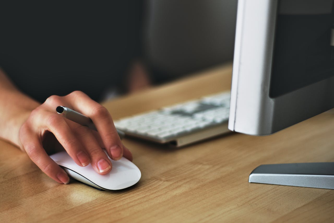 Hand using a wireless mouse next to a desktop monitor and keyboard on wooden desk