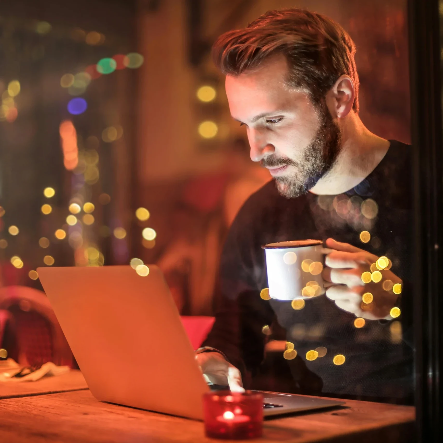 Man working on a laptop in a cozy café with ambient lighting, holding a coffee mug