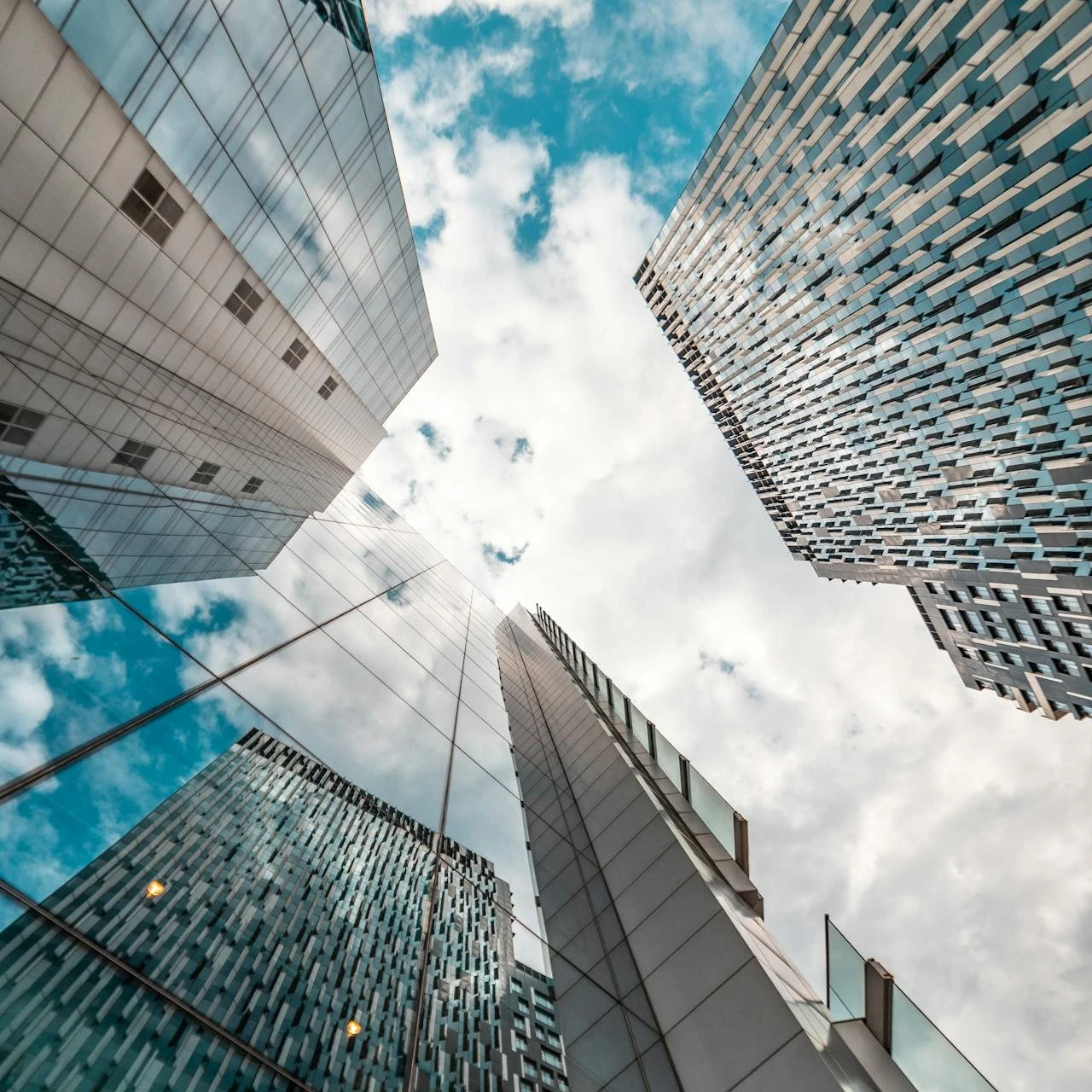Upward view of modern glass skyscrapers under a cloudy blue sky, reflecting contemporary architecture and ambition
